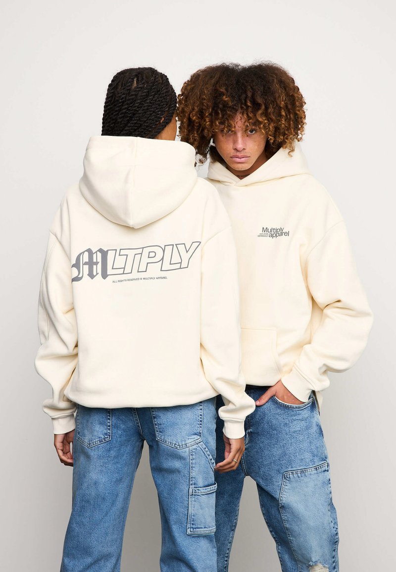 Crème-kleurige hoodies hebben een groot grijs "MLTPY" logo op de achterkant en een kleiner logo op de voorkant. Gemaakt met blauwe cargobroeken.