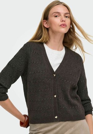 Femme aux longs cheveux blonds portant un cardigan en maille foncé avec des boutons en forme de cœur sur une chemise blanche, posant devant un fond neutre.