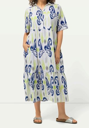 Vrouw draagt een midi-jurk met wit, blauw en limoengroen patroon, korte mouwen en knopen aan de voorkant, gecombineerd met zilveren sandalen met open tenen.