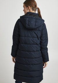 Manteau rembourré bleu marine avec capuche, présentant un design matelassé horizontal, un tissu texturé et des poignets élastiques ; arrive à mi-mollet.