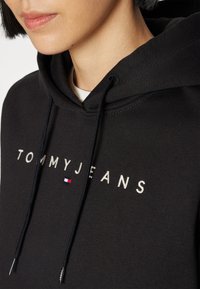 Sudadera negra con el bordado "TOMMYJEANS" en letras blancas y un pequeño logo en rojo, blanco y azul. Cuenta con cordones y una textura suave.