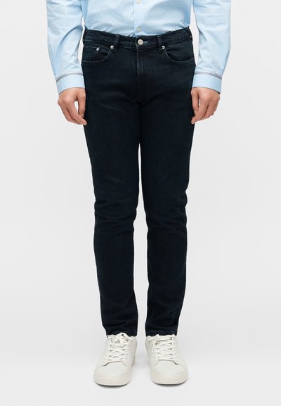 Jean en denim foncé, coupe slim, avec un design classique à cinq poches et des coutures contrastantes, associé à des baskets blanches.