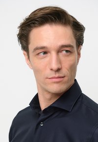 Donker marineblauw overhemd met knopen, een kraag, een gladde textuur en een enkele knoopdetail. Het model heeft kort, rommelig haar en een neutrale uitdrukking.