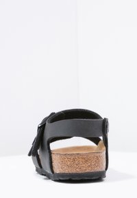 Musta nahkainen sandaali, jossa säädettävä remmi, korkkipohjallinen, kuvioitu ulkopohja ja takaremmin muotoilu. Varustettu pyöreällä napakiinnityksellä.