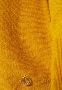 Détail d'un pull en tricot jaune avec une texture côtelée, accompagné d'un pendentif en forme de cœur argenté accentuant l'avant.