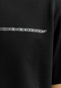 Sort bomulds t-shirt med en grå vandret stribe, hvor teksten "JACK & JONES" står i hvid. Korte ærmer og glat tekstur.