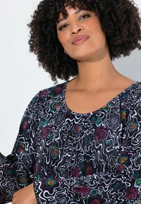 Schwarzes, plissiertes Oberteil mit einem wellenförmigen weißen Linienmuster und bunten floralen Akzenten, Rundhalsausschnitt und langen Ärmeln. Weiche Stofftextur.