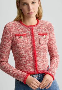 Cardigan lavorato a maglia rosso e bianco con finitura testurizzata, bordi frangiati, due tasche sul petto e dettagli in bottone dorato. Maniche lunghe, design aderente.