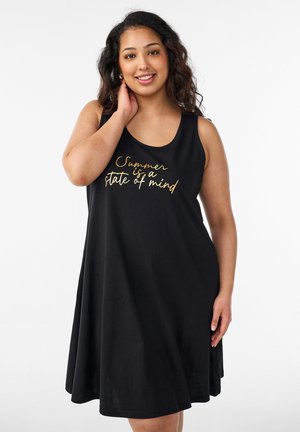 Mujer sonriente con cabello ondulado lleva un vestido negro sin mangas con texto dorado «El verano es un estado mental» sobre un fondo blanco.