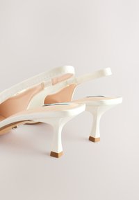 Escarpins slingback blancs à talons fins avec embouts en caoutchouc beige sur un fond clair.