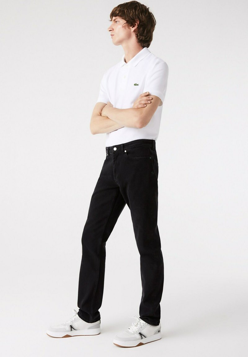 Schwarze Jeans mit slim fit, kombiniert mit einem weißen Poloshirt mit einem kleinen grünen Logo. Die Schuhe sind weiß mit grauen Akzenten.