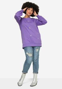 Sudadera morada de manga larga, adornada con detalles plateados, combinada con unos vaqueros desgastados que tienen detalles de perlas, y botas de tobillo plateadas.