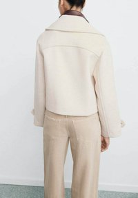 Beżowa kurtka z wełny z kołnierzowym dekoltem, krótkim krojem i długimi rękawami. Połączona z beżowymi spodniami z szerokimi nogawkami, posiadającymi tylnie kieszenie.