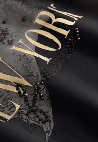 Zwarte stof met gouden letters en zwarte pailletten in een decoratief patroon, met een gestructureerde afwerking en subtiele grafische details.