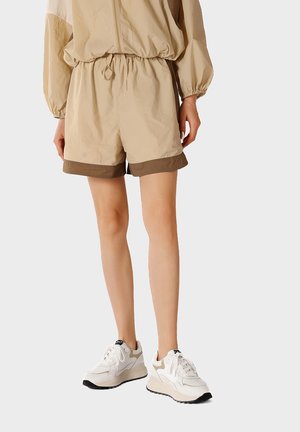 Modella che indossa pantaloncini beige a vita elastica con rifiniture marroni, top beige a maniche lunghe e sneakers bianche chunky, in piedi su uno sfondo chiaro.