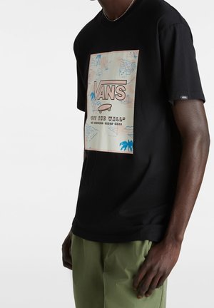 Personne portant un t-shirt noir Vans avec un logo skateboard et le texte « Off The Wall », associé à un pantalon vert clair.