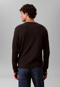 Brązowa koszula z długim rękawem o fakturowanym, prążkowanym wzorze, zestawiona z niebieskimi dżinsami. Koszula ma okrągły dekolt i lekko luźny krój.