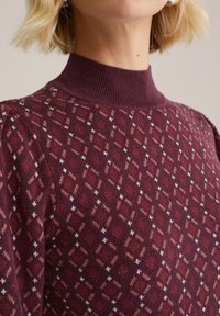 Bordeauxkleurige trui met een geribde halslijn, voorzien van een diamantpatroon in verschillende tinten roze en wit over het oppervlak van de stof.
