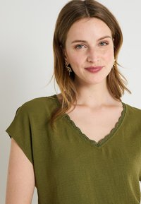 Blouse vert olive en tissu texturé. Présente un décolleté en V festonné et des manches courtes. Le design met en avant une coupe décontractée.