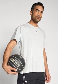 Αθλητική μπλούζα ανοιχτού γκρι χρώματος με κοντά μανίκια και βαθμιδωτό μοτίβο, φορεμένη με μαύρο σορτς, κρατώντας μια μπάλα μπάσκετ στο χέρι.