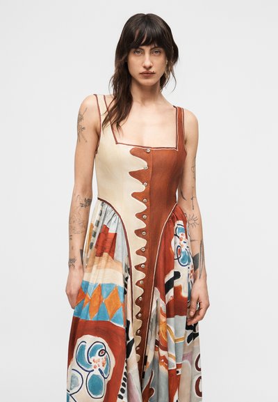 Γυναίκα με σκούρα μαλλιά, που φοράει αμάνικο πολύχρωμο φόρεμα με κουμπιά και αφηρημένα σχέδια, στέκεται μπροστά σε απλό φόντο.