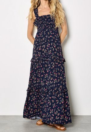 Femme portant une robe maxi fleurie bleu marine avec volants et bretelles réglables, associée à des sandales plates couleur beige, debout contre un mur uni.