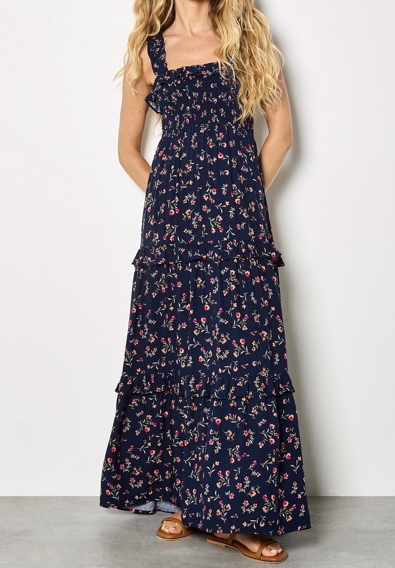 Femme portant une robe maxi fleurie bleu marine avec volants et bretelles réglables, associée à des sandales plates couleur beige, debout contre un mur uni.