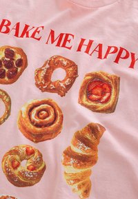 Roze T-shirt met verschillende gebakafbeeldingen, waaronder kaneelrollen, croissants en donuts, met de tekst "BAKE ME HAPPY" in vetgedrukte rode letters.
