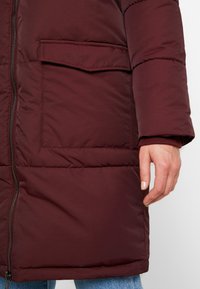 Cappotto imbottito bordeaux con chiusura a zip, dotato di tasca laterale e polsini a costine. Tessuto liscio con una leggera lucentezza.