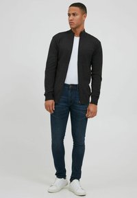 Mörkgrå zip-up-jacka över vit skjorta, i kombination med mörkblå jeans och vita sneakers. Har en uppstående krage och en figurnära design.