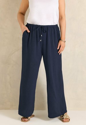 Mujer con pantalones de pierna ancha azul marino con cintura ajustable, top blanco sin mangas, pulseras doradas y sandalias doradas, de pie contra una pared beige.