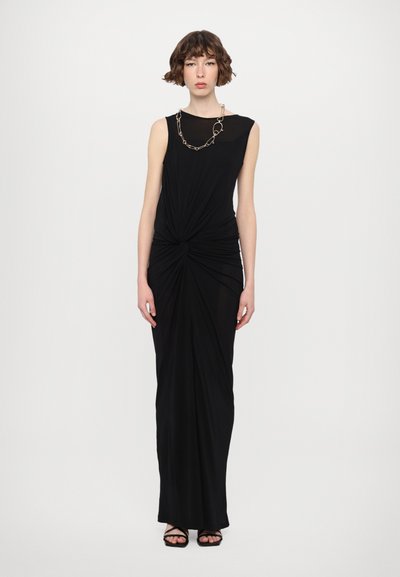 Vestido negro con tela drapeada y un frente retorcido. Presenta un escote alto y un collar decorativo de cadena. Se lleva con sandalias negras.