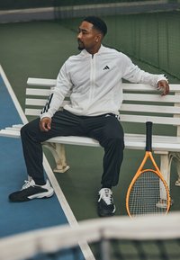 Adidas witte ritsjack met zwarte strepen, zwarte sportieve broek en zwart/witte sneakers. Een oranje tennisracket ligt op een bank.