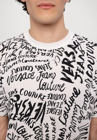 Balta kokvilnas T-kreklā ir haotiska melna grafika ar "Versace Jeans Couture" dažādās kursīvā fonta stilā un orientācijās.