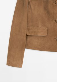 Giacca in suede marrone chiaro con chiusura a bottone, una tasca e superficie liscia, caratterizzata da una vestibilità sartoriale e taglio dritto.