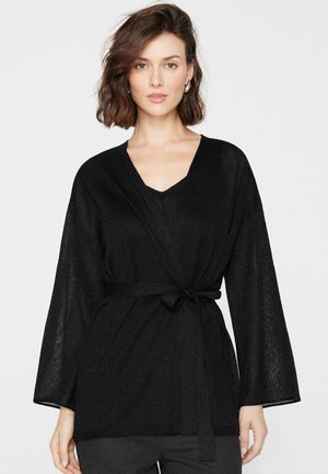 Donna con capelli castani fino alle spalle che indossa un maglione nero incrociato con scollo a V annodato in vita, in piedi davanti a uno sfondo bianco semplice.