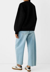 Svart oversized sweatshirt med sänkta axlar, kombinerad med ljusblå vida jeans med fransade kanter, och vita sneakers med svarta accenter.