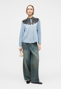 Γυναίκα που φοράει μπλε ριγέ μπλούζα με μαύρο δαντελένιο γιακά, φαρδιά ξεβαμμένα τζιν, μαύρα παπούτσια και κρατάει μια μικρή μπεζ τσάντα.