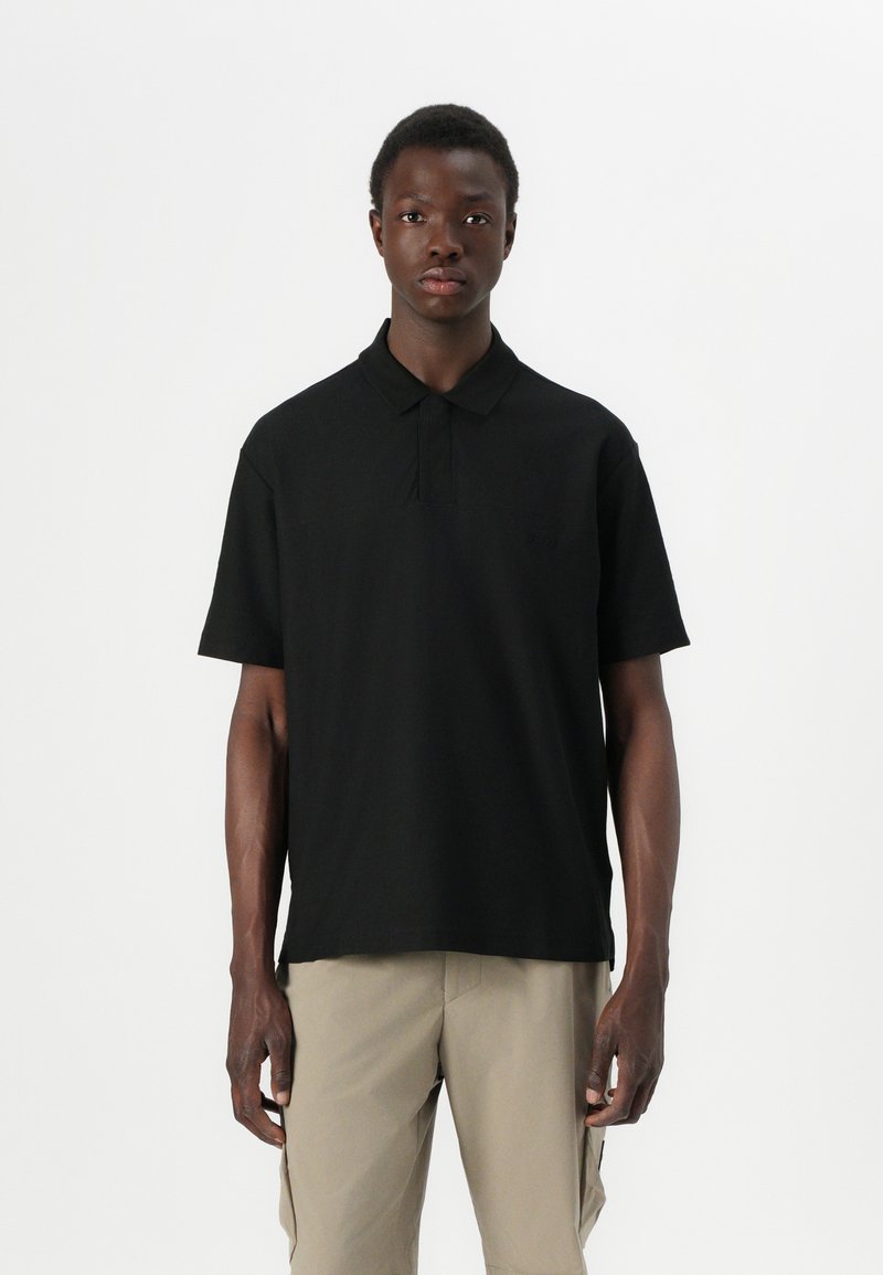 Camisa polo preta em um corte descontraído, com colarinho clássico, mangas curtas e detalhes de costura sutis. Usada com calções claros em caqui.