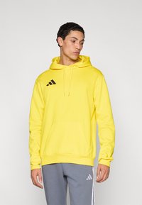 Κίτρινο φούτερ με κουκούλα, τσέπη στο μπροστινό μέρος, κορδόνι κουκούλας και μαύρο λογότυπο Adidas. Συνδυασμένο με γκρι φόρμα με λευκές ρίγες.