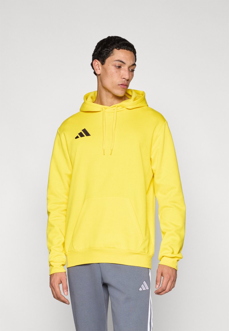Κίτρινο φούτερ με κουκούλα, τσέπη στο μπροστινό μέρος, κορδόνι κουκούλας και μαύρο λογότυπο Adidas. Συνδυασμένο με γκρι φόρμα με λευκές ρίγες.