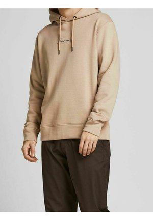 Beiger Hoodie mit Kordelzug und kleinem Schriftzug-Logo auf der Brust, getragen von einer stehenden Person vor einfachem Hintergrund, kombiniert mit dunkelbraunen Hosen.