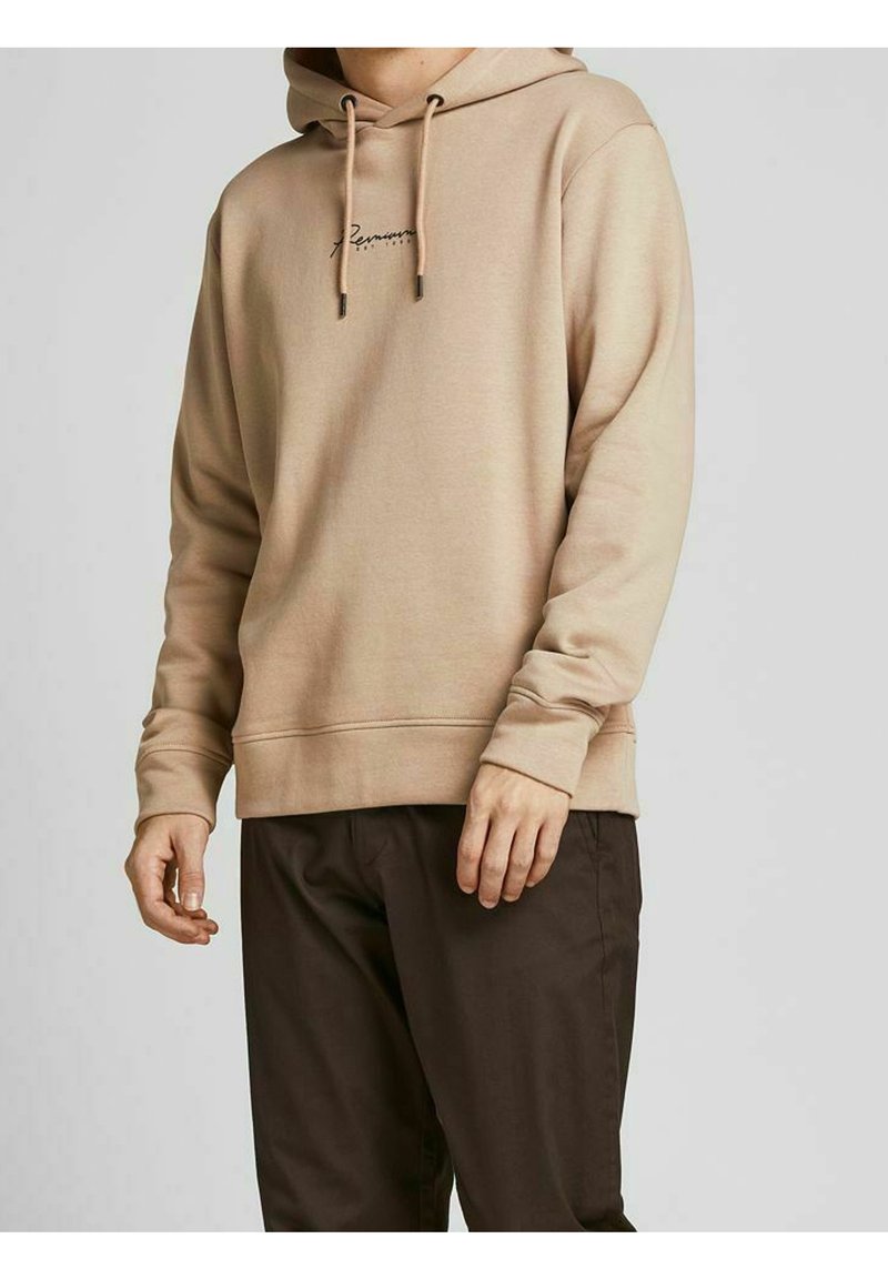 Beige hættetrøje med snore og lille skrevet logo på brystet båret af en stående person iført mørkebrune bukser mod en ensfarvet baggrund.