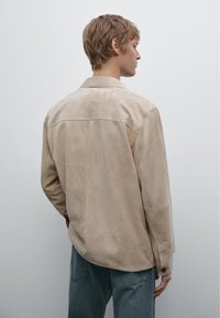 Beige suede jacket with a classic collar, long sleeves, and buttoned cuffs. The back features a yoke seam and smooth texture.
