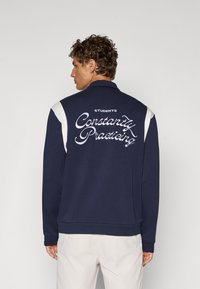 Marineblå hettegenser med hvite skuldrekanter og brodert tekst som sier "STUDENTER Øver Konstant" på ryggen. Glatt stofftekstur.