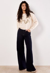 Un pull crème avec du texte doré pailleté, associé à un jean bleu foncé à jambes larges. Les deux vêtements présentent des textures douces et des designs décontractés.