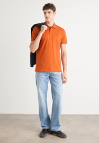 Oransje polo med krage, korte ermer og en liten logo, paret med lyseblå jeans og svarte joggesko.