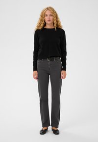 Čierny sveter, šedé rovné nohavice s opaskom, čierne ploché topánky. Pozadie je jednoduché biele, zameranie je na oblečenie a model.