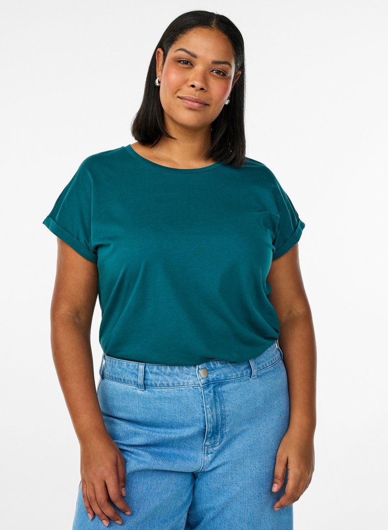 Donna con capelli castani scuri di media lunghezza che indossa una maglietta a maniche corte color verde acqua e jeans azzurri a vita alta su uno sfondo bianco semplice.