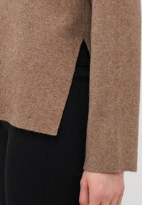 Brauner gerippter Wollpullover mit seitlichen Schlitzen, langen Ärmeln und einer entspannten Passform, kombiniert mit schwarzen Hosen für einen Kontrast.